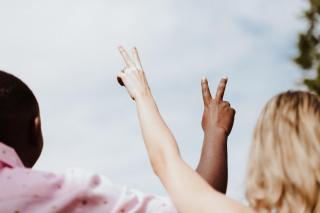To people--one white and one Black--hold up their arms, each hand forming the "peace" symbol of two fingers in a V.