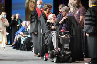 A line of ministers cross the stage to be recognized by officiants. The foreground focuses on Rev. Clyde Grubbs, a religious professional completing service, being greeted by Dr. Janice Marie Johnson, co-director of the UUA Office of Ministries and Faith Development..