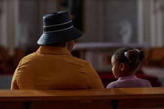 From the back, a Black woman wearing a hat and a small Black child are seated in a church pew.