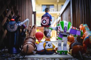 A full range of dolls from the Toy Story movie series is arranged on a porch, backs to the viewer, watching a small child wearing a backpack leave the house 