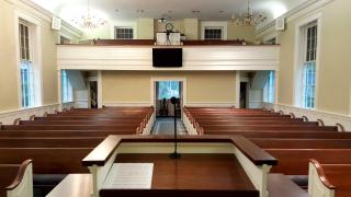 A view of wooden church pews, and the church narthex, from the pulpit. The pulpit holds a piece of sheet music and a microphone.