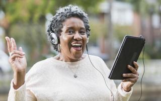 A Black woman wearing headphones smiles and sings, holding a tablet or iPad.