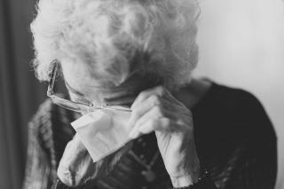 An elderly person with short curly white hair adjusts their glasses so they can wipe away tears with a tissue.
