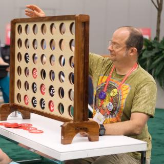 A man wearing a green shirt sits at white table playing a large connect-four game with red and black pieces. 
