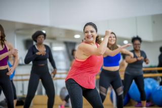 In a dance studio, a woman in a red tank top dances ethusiastically while other dancers are visible in the background.