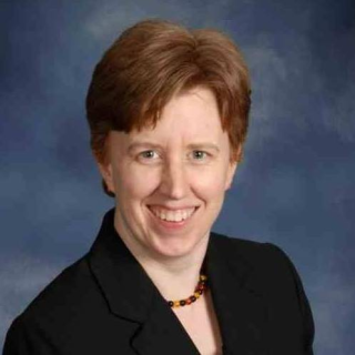 Erica Baron: Head and shoulders of a white woman with short red hair, wearing a black blazer and a black and orange necklace against a mottled blue background.