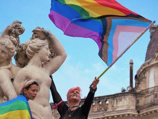 A woman waves a progress pride flag (a rainbow flag with additional concentric triangles in white, pink, blue, brown, and black) while standing near the top of a statue of the Three Graces in Montpellier, France. A second person, wrapped in a pride flag, stands nearby on the statue. 