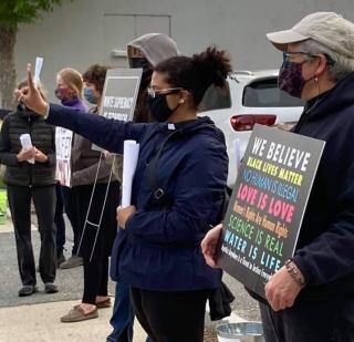 Vanessa, a Black person with her hair in a ponytail, stands between church members with signs. Vanessa is wearing a mask and clergy collar, and is holding her hand in the "peace" sign.