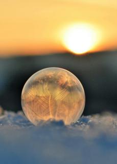 A frozen soap bubble rests on snowy ground, with the sun setting on the horizon in the background.