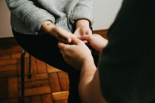 A cropped photo of a person with a wrist tattoo with upturned hands lightly holding the hands of another person, sitting knee-to-knee with them.