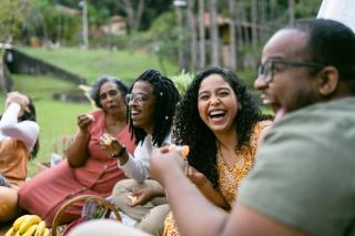 Outdoors on a lawn, a group of Black friends are seated as if at a picnic. They're smiling and laughing with abundant joy.