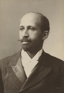 Formal portrait photograph of W. E. B. Du Bois, a balding Black man with a mustache and Van Dyke beard, wearing a white shirt and dark coat.