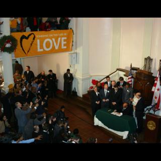 People gather around Washington, DC, Mayor Adrian Fenty as he signs a bill legalizing equal marriage. A 'Standing on the Side of Love' banner hangs over the balcony above.