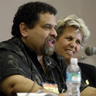 Jos&eacute; Ballester, foreground, speaking into a microphone; Kim Crawford Harvie, background, smiling.