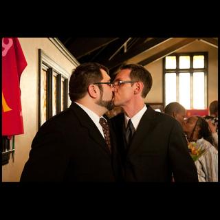 A pair of men in suits share a kiss in a church, other couples behind them also sharing an embrace or kiss.