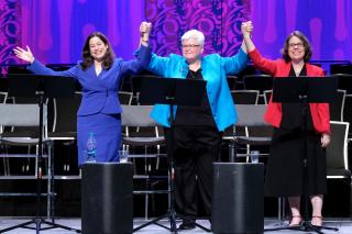 The Rev. Alison Miller, the Rev. Jeanne Pupke, and the Rev. Susan Frederick-Gray stand on stage, arms raised and hands clasped.