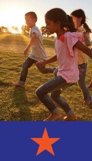 three grade school age kids in jeans and summer tops run barefoot across a field with trees' long shadows in background