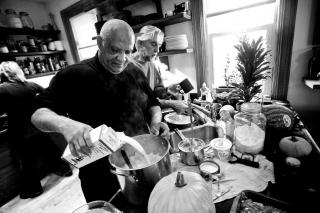 In a black and white photo, someone pours milk into a large pot of mashed potatos. Behind him, someone else prepares food at a kitchen counter.