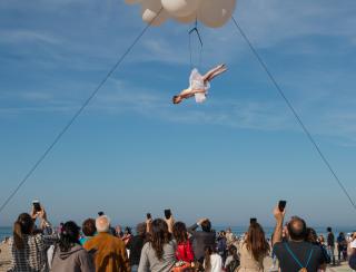 Above a crowd of people looking up and taking pictures with their phones, a white-tutu-wearing trapeze artist is suspended from a cloud of white balloons (part of the Molecole show)..