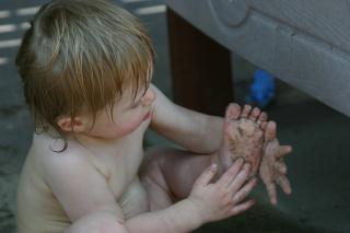 A baby plays with her own foot, with hands and foot covered in sand or dirt