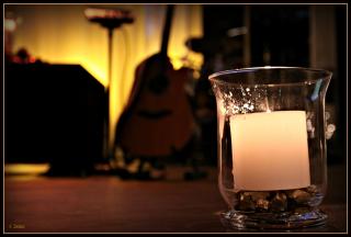 An ivory pillar candle in a clear hurrican holder, with blurred musical instruments in the background.