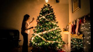 A child stands, putting the final touches on a lit Christmas tree in a dark room.