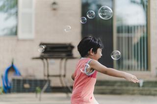 A small child, seen in profile with a huge smile on their face, runs through soap bubbles in a back yard.
