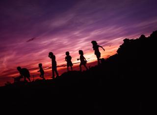 A line of children (and a dog) in silhouette against the purples and oranges of sunset.