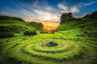 The sun rises between two green hills. In the foreground, circles of stone and earth radiate outward from a stone center.