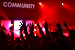 In a gathering lit by red lights, the word COMMUNITY appears on the wall as many arms wave in the air.