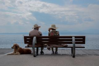 An elderly couple, wearing hats, sits on a bench with their backs to the camera. A dog lounges at their feet. 