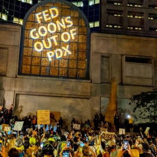 Image of crowd of protesters in Portland, OR, where the words "Fed goons out of PDX" are projected on wall