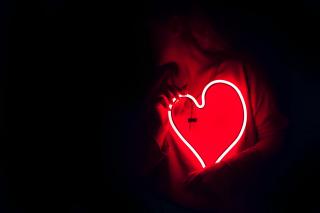 A person, seen only from the waist up in the dark, holds a red neon heart in their hands.