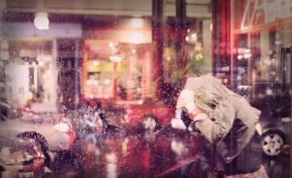Through a plate glass window at night, a person ducks through rainy weather with their coat pulled over their head.