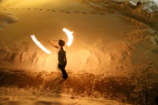 Seen from above, in light cast only by flames, a solitary person swings two "poi" fire weights on chains (sometimes called "fire juggling")