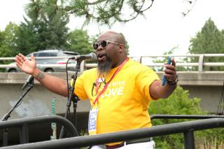 An African American man in a bright yellow "Side with Love" t-shirt speaks at a microphone and gestures broadly with his hands