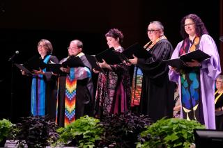 Five worship leaders in colorful stoles and holding large black folders appear on stage during the worship