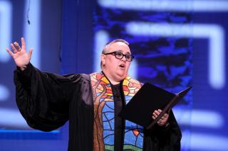 A preacher in black robes and a stained glass pattern stole gestures during a sermon.