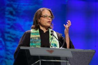 An African American preacher with a green stole gestures from the podium