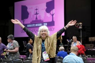An exuberant woman, wearing a shawl, spreads her arms in celebration and welcome. A projection of the pink GA logo forms a backdrop.