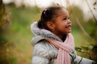 A young girl, bundled into a warm coat, smiles excitedly at someone off-camera.