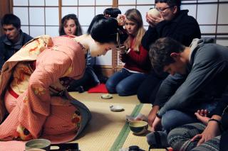 A kneeling Geisha and a white male in western clothing bow to each other in a tea ceremony, as westerners drink tea in the background.