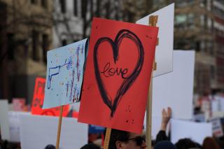 A sea of signs, above people's heads at a rally or protest. The center sign on red posterboard is a large black heart with "love" written inside.