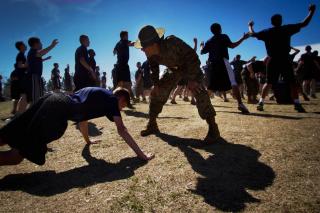 Amidst a field of men doing jumping jacks, a drill instructor in uniform bends over to shout at a man doing push-ups.