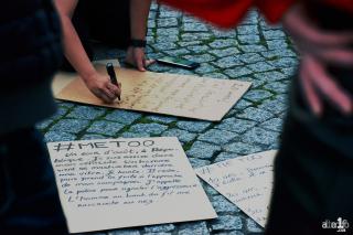 On a cobblestone surface, disembodied hands write signs with #METOO at the top.