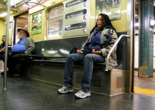 On a NYC subway car, a woman sits with both feet planted on the floor, hands held lightly in meditation position, with her eyes closed.