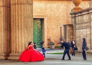 A team of professional photographers, outdoors, takes photos of a quinceañera in a billowing red formal gown.