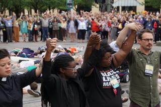 People stand in the in a circle holding upraised hands with others lay on the ground in the center.