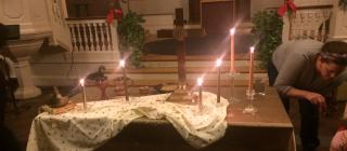 a table covered in decorative fabric holds 6 candles and a cross, in the background you can see Christmas decorations and more candles