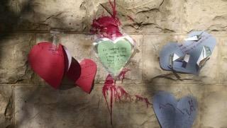 Heart-shaped notes are taped to a wall where paint has been thrown.